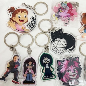 May include: A collection of acrylic keychains with various designs. Keychains include cartoon characters, a photo of a person, and a black cat on a die. The keychains have silver metal rings.