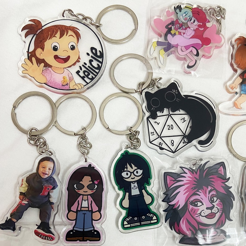 May include: A collection of acrylic keychains with various designs. Keychains include cartoon characters, a photo of a person, and a black cat on a die. The keychains have silver metal rings.