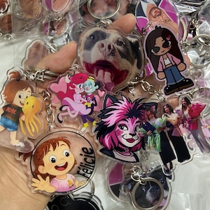 May include: A collection of clear acrylic keychains featuring various designs. The keychains showcase cartoon characters, a dog's face, and images of people. Each keychain has a silver metal ring for attachment.