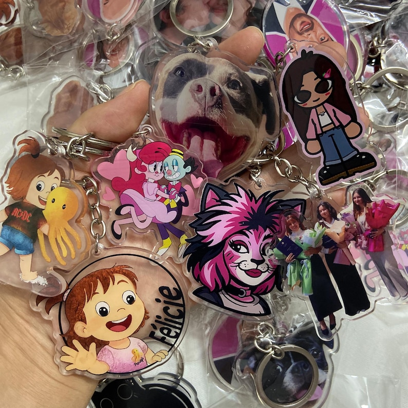 May include: A collection of clear acrylic keychains featuring various designs. The keychains showcase cartoon characters, a dog's face, and images of people. Each keychain has a silver metal ring for attachment.