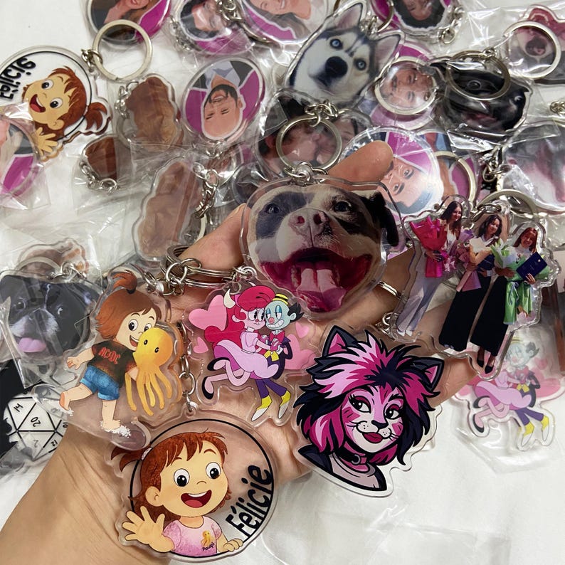 May include: A collection of clear acrylic keychains with various images. The keychains feature images of dogs, cartoon characters, and people. Some keychains have text, including the name "F&eacute;licie". The keychains are attached to silver metal keyrings.