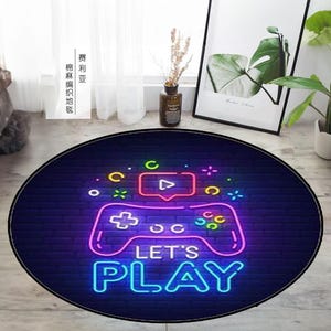 Puede incluir: Una alfombra redonda de color azul oscuro con un mando de videojuego de color rosa neón, azul, verde y amarillo y las palabras "Let's Play" en una fuente de neón.