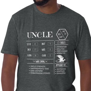 May include: A dark heather grey t-shirt with a white graphic of a character sheet for a game. The graphic says "Uncle" and has stats for strength, intelligence, dexterity, wisdom, constitution, and charisma. The graphic also includes skills, immunities, and weaknesses.