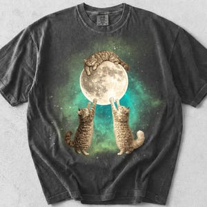 Könnte beinhalten: Dunkelgraues T-Shirt mit einem fantasievollen Design von drei Katzen, die mit einem Mond interagieren, wobei eine Katze obenauf ruht. Die Grafik ist vor dem Hintergrund eines sternenklaren, türkis-grünen Nebels angeordnet. Das Wort "Pepper" ist unten aufgedruckt.