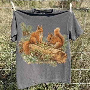 May include: A gray t-shirt featuring a detailed illustration of two squirrels on a log with pine cones and foliage. The shirt is hanging on a clothesline against a backdrop of a field and blue sky.
