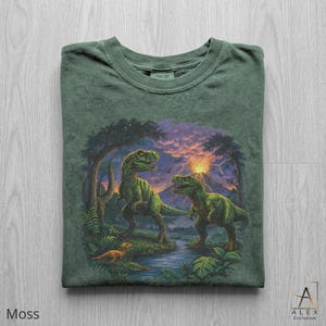May include: Moss green t-shirt featuring a detailed illustration of two green dinosaurs in a prehistoric landscape with a volcano and sunset. The shirt has a crew neck and the word "COMFORT" is printed on the tag.