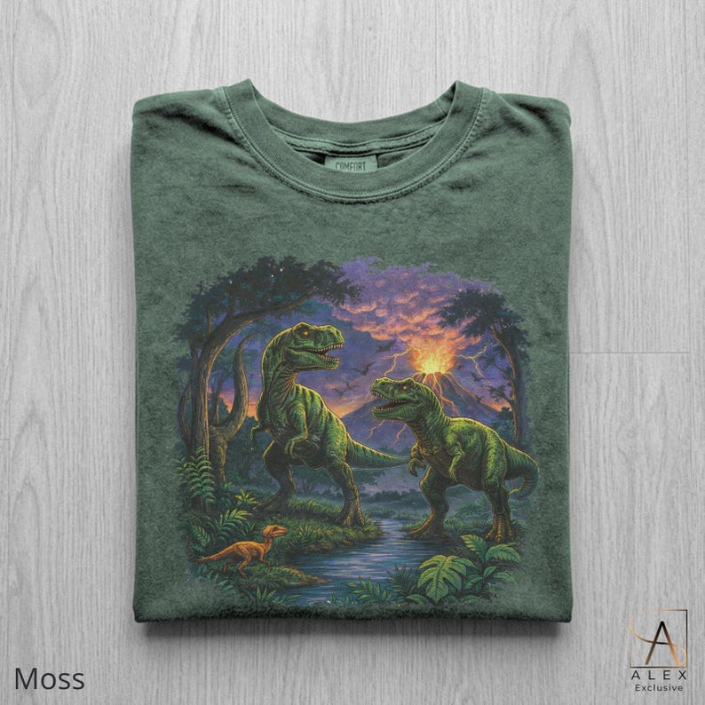 May include: Moss green t-shirt featuring a detailed illustration of two green dinosaurs in a prehistoric landscape with a volcano and sunset. The shirt has a crew neck and the word "COMFORT" is printed on the tag.