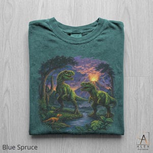 May include: Blue spruce t-shirt featuring a detailed illustration of two green dinosaurs in a prehistoric landscape with a river, trees, and a volcano. The shirt has the word "COMFORT" printed on the collar. The text "Blue Spruce" is printed at the bottom.