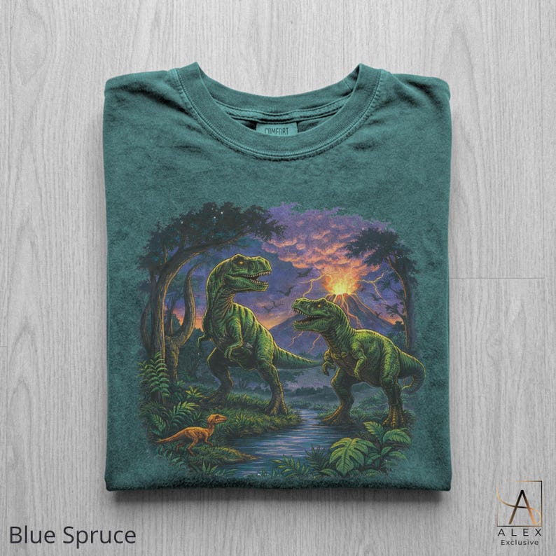 May include: Blue spruce t-shirt featuring a detailed illustration of two green dinosaurs in a prehistoric landscape with a river, trees, and a volcano. The shirt has the word "COMFORT" printed on the collar. The text "Blue Spruce" is printed at the bottom.