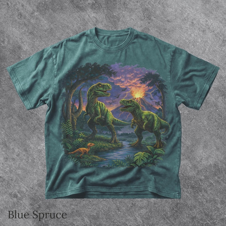 May include: Blue Spruce t-shirt featuring a detailed illustration of two green Tyrannosaurus Rex dinosaurs in a prehistoric landscape with a river, trees, and a volcano. The shirt has the text "Blue Spruce" at the bottom.