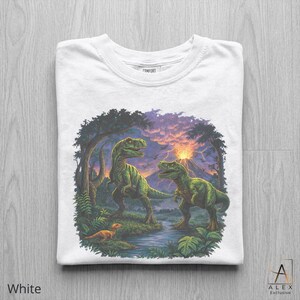 May include: White t-shirt featuring a detailed illustration of two green dinosaurs in a prehistoric landscape with a volcano and a small dinosaur. The shirt has the word "COMFORT" printed on the collar and the words "ALEX Exclusive" on the bottom right.