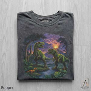 May include: Pepper-colored t-shirt featuring a vibrant dinosaur scene. Two green dinosaurs stand near a river, with a smaller dinosaur in the foreground. The background includes a volcano, trees, and a colorful sunset. The shirt has a crew neck.