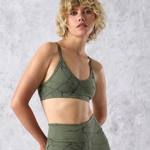 Peut inclure: Ensemble de soutien-gorge de sport et de legging vert olive avec un motif marbré noir. Le soutien-gorge a de fines bretelles et une encolure en forme de cœur. Le legging est taille haute. Le modèle porte l'ensemble devant un fond gris.