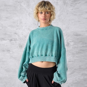 Pode incluir: Uma camisola curta turquesa com mangas compridas e gola redonda. A peça de roupa tem um aspeto desbotado e vintage. A modelo usa calças pretas de cintura alta. O fundo é cinzento e branco salpicado.