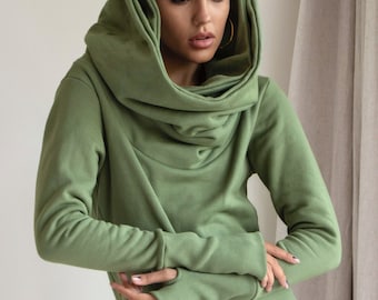 May include: A long-sleeved, green hooded dress with a cowl neck and thumbholes. The dress has a relaxed fit and a slightly asymmetrical hemline.