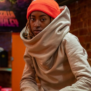 Könnte beinhalten: Eine Person trägt einen beigen Kapuzenpullover mit einem großen, gefalteten Kragen. Die Person trägt eine leuchtend orangefarbene Mütze und hat Dreadlocks. Die Person sitzt mit verschränkten Armen und blickt in die Kamera.