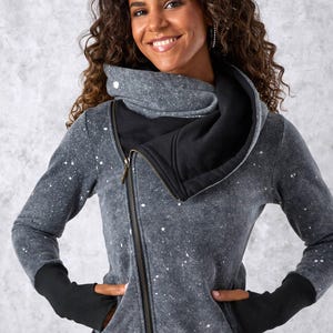 Puede incluir: Una sudadera con capucha y cremallera en gris carbón con un patrón de salpicaduras de pintura blanca, un cuello asimétrico de gran tamaño, mangas largas con puños negros con orificios para los pulgares y un dobladillo negro. Cremallera diagonal.