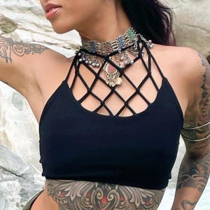 May include: A woman with dark hair and tattoos wears a black halter top with a mesh overlay and a silver and turquoise choker necklace. She is wearing a silver headpiece with dangling charms.
