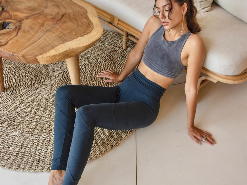 Op de afbeelding: Een vrouw in een donkergrijs tanktop en donkerblauwe leggings poseert op een geweven tapijt.
