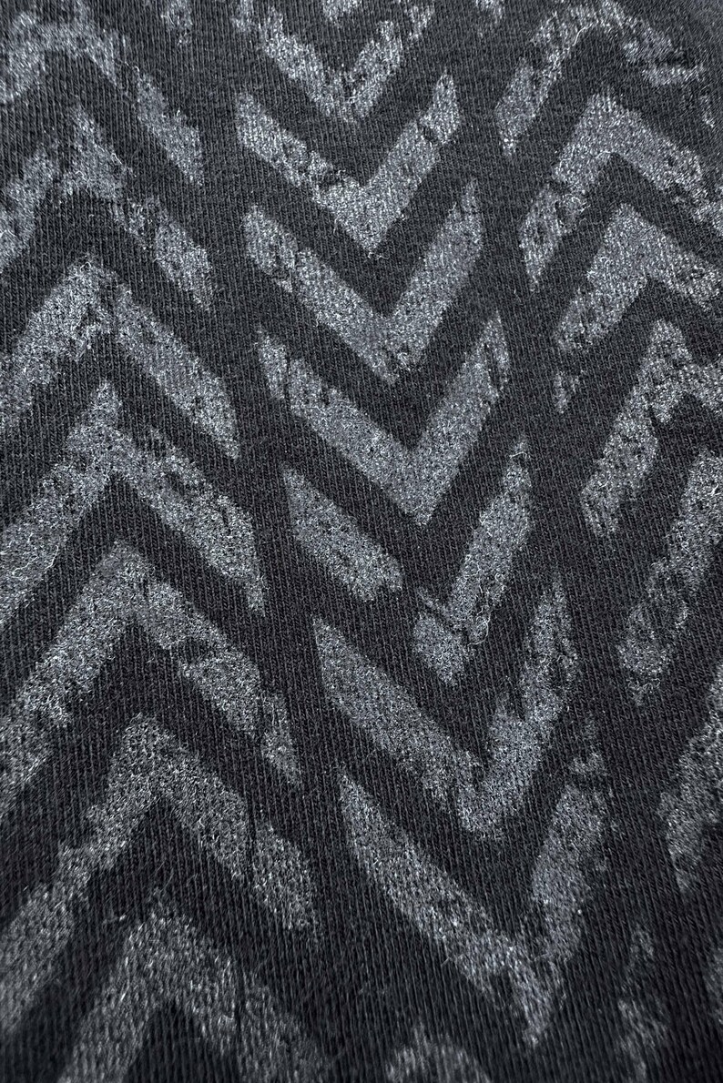 May include: Close-up of a black fabric with a repeating chevron pattern in shades of gray. The design has a textured, distressed appearance, creating a visual contrast between the dark background and the lighter pattern.