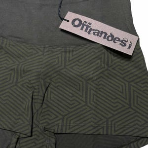May include: Olive green athletic shorts with a geometric pattern in a darker shade of green. The shorts have a high waistband and a tag that reads "Offrandes".