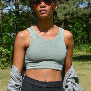 Op de afbeelding: Een vrouw draagt een saliegroen getailleerd tanktop met een zwarte broek met hoge taille en een grijs shirt dat over haar schouders is gegooid.