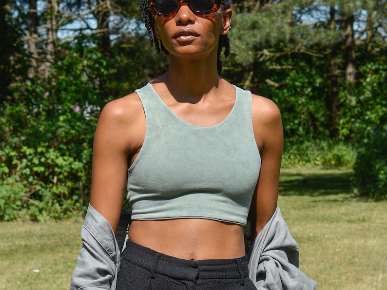 Op de afbeelding: Een vrouw draagt een saliegroen getailleerd tanktop met een zwarte broek met hoge taille en een grijs shirt dat over haar schouders is gegooid.