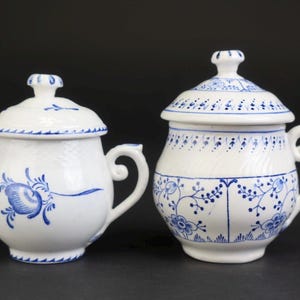Op de afbeelding: Twee witte keramische potten met deksel en blauwe bloemmotieven. Elke pot heeft een bolvormig lichaam, een gebogen handvat en een bijpassend deksel met een kleine knop. De linkerpot heeft een enkel groot bloemmotief op de zijkant.