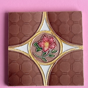 May include: A square ceramic tile with a brown background and a white and yellow geometric design. A pink flower with green leaves is in the centre of the tile.