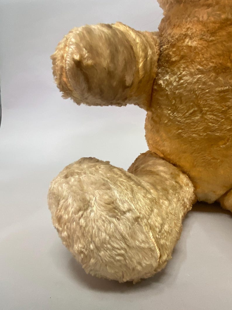May include: Close-up of a vintage teddy bear's feet and arm. The bear is made of a soft, light brown plush fabric. The feet are slightly worn and have a faded appearance.