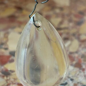 May include: A clear, teardrop-shaped crystal pendant with a small hole at the top, suspended by a dark wire. The crystal reflects light, and is attached to a chain with other crystals.