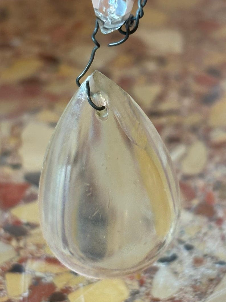May include: A clear, teardrop-shaped crystal pendant with a small hole at the top, suspended by a dark wire. The crystal reflects light, and is attached to a chain with other crystals.