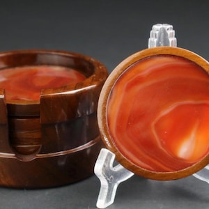 May include: A set of four coasters with a holder. The coasters have a reddish-orange and white marbled design set in a wooden frame. The holder is made of the same dark brown wood and has a slot for each coaster.