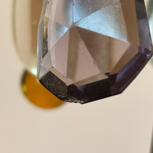 May include: Close-up of a faceted, teardrop-shaped crystal pendant. The crystal is a smoky, translucent color with a geometric design. The edges of the crystal are slightly chipped. The background is blurred, with hints of gold and white.