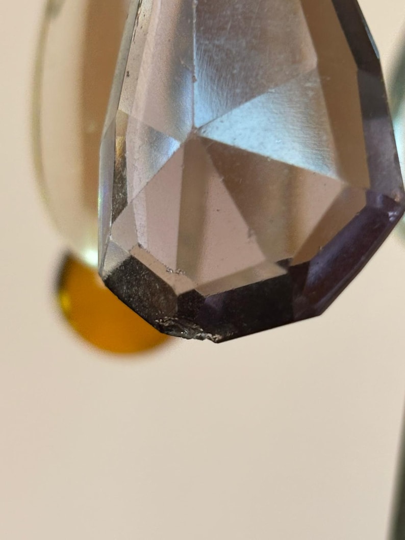 May include: Close-up of a faceted, teardrop-shaped crystal pendant. The crystal is a smoky, translucent color with a geometric design. The edges of the crystal are slightly chipped. The background is blurred, with hints of gold and white.