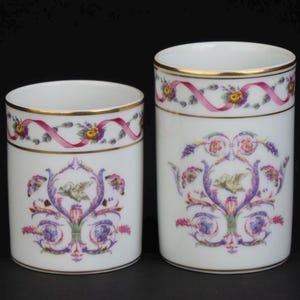 May include: Two white porcelain cylindrical containers of different heights, each featuring a gold rim and a decorative band of pink ribbons, yellow flowers, and green leaves, with a central motif of a bird surrounded by purple and pink scrollwork.