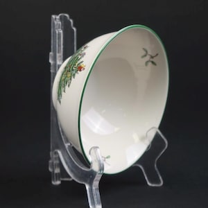 May include: A cream-coloured ceramic bowl with a thin green rim and a festive Christmas tree graphic on the exterior. The bowl is displayed on a clear acrylic stand, showcasing a small sprig of mistletoe painted on the interior surface.