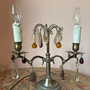 May include: A vintage candelabra-style lamp with two faux candle lights. The lamp features a silver-toned base and arms, adorned with clear, amber, and purple crystal accents. The lamp has a power cord and is designed to sit on a table.