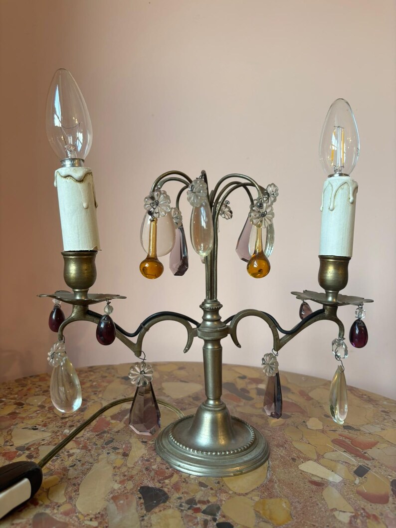 May include: A vintage candelabra-style lamp with two faux candle lights. The lamp features a silver-toned base and arms, adorned with clear, amber, and purple crystal accents. The lamp has a power cord and is designed to sit on a table.