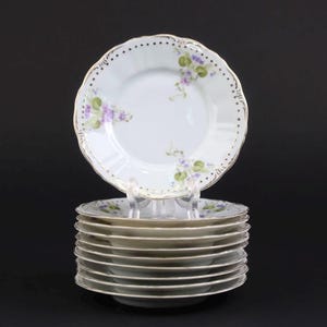 Könnte beinhalten: Ein Stapel weißer Porzellanteller mit gewelltem Rand und Goldverzierung. Jeder Teller zeigt ein zartes Blumenmuster aus kleinen violetten Blüten und grünen Blättern, akzentuiert durch einen Ring aus winzigen goldenen Punkten am Rand.