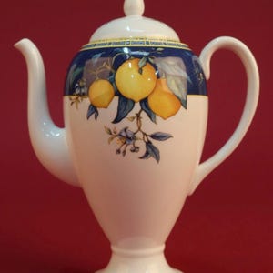 May include: White ceramic teapot with a lid, featuring a blue band with yellow lemons and green leaves. The teapot has a curved spout and handle, and sits on a pedestal base. The background is a solid red.