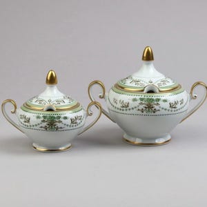 Może przedstawiać: Dwie białe cukiernice porcelanowe z pokrywkami, posiadające złocone uchwyty, sterczyny i podstawy. Naczynia ozdobione są wzorem zielono-złotych girland kwiatowych, misternymi zawijasami oraz kropkowanym obramowaniem wokół środkowej części.