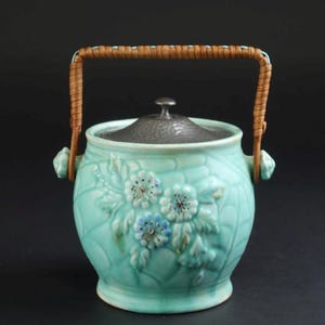 Puede incluir: Un tarro de cerámica de color turquesa claro con un patrón floral en relieve texturizado en la parte frontal. El tarro cuenta con una tapa de metal martillado gris oscuro y un asa curva de mimbre trenzado marrón claro en los laterales.