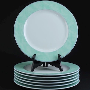 Può includere: Una pila di sei piatti piani rotondi con un centro bianco solido e un ampio bordo verde menta marmorizzato. Il piatto superiore è tenuto in posizione verticale da un supporto in plastica nera, che mette in risalto la finitura liscia.