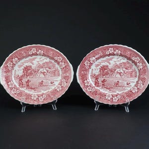 May include: A pair of oval ceramic platters featuring a red and white transferware pattern. The design depicts a horse-drawn carriage scene with figures and dogs, surrounded by a decorative floral border with a scalloped edge on each plate.