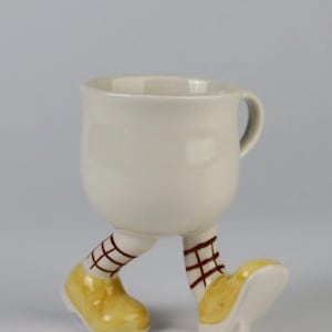 Puede incluir: Una taza de cerámica caprichosa en blanco roto, con piernas con zapatos amarillos y calcetines marrones con entramado. La taza tiene un asa y un cuerpo redondeado, creando un diseño lúdico. Una taza original para la cocina.