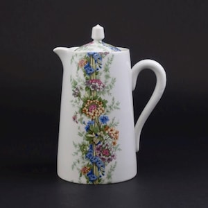 May include: A white porcelain coffee pot featuring a vertical floral pattern of blue, pink, yellow, and green flowers arranged along a central stem. The pot has a curved handle, a spout, and a matching lid with a small, pointed finial on top.