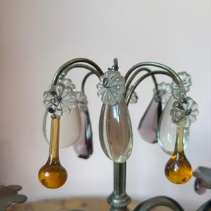 May include: Close-up of a decorative chandelier with a silver metal frame. The chandelier features clear, teardrop-shaped crystal pendants in various colors, including amber and purple. The design includes flower-shaped accents.