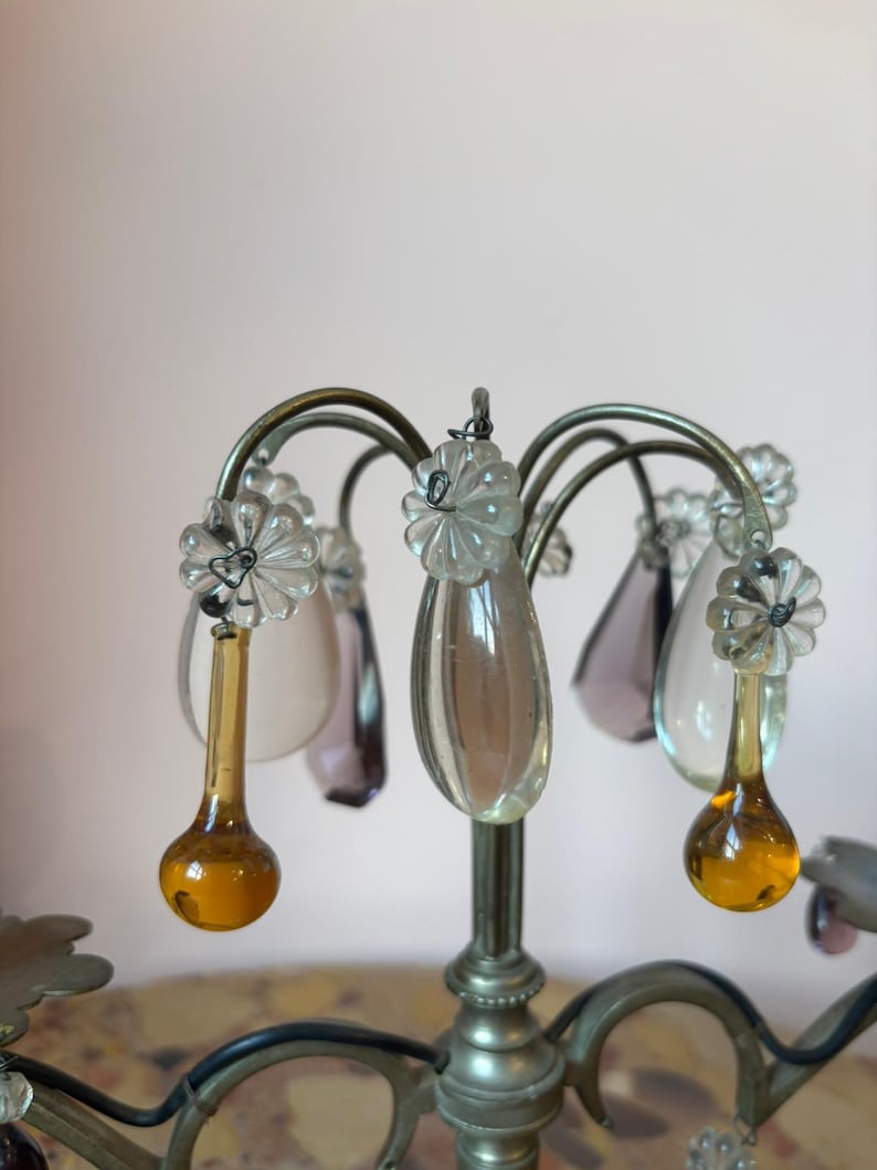 May include: Close-up of a decorative chandelier with a silver metal frame. The chandelier features clear, teardrop-shaped crystal pendants in various colors, including amber and purple. The design includes flower-shaped accents.