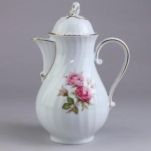 Puede incluir: Una cafetera de porcelana blanca con textura acanalada vertical y un delicado patrón floral de rosas rosadas con hojas verdes. La cafetera incluye una tapa a juego con un remate en forma de capullo, un asa curva y bordes con detalles dorados.